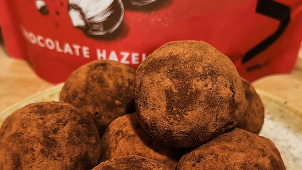 Dark chocolate protein balls dusted with cocoa, made without nut butter, in front of a WAM Chocolate Hazelnut pouch