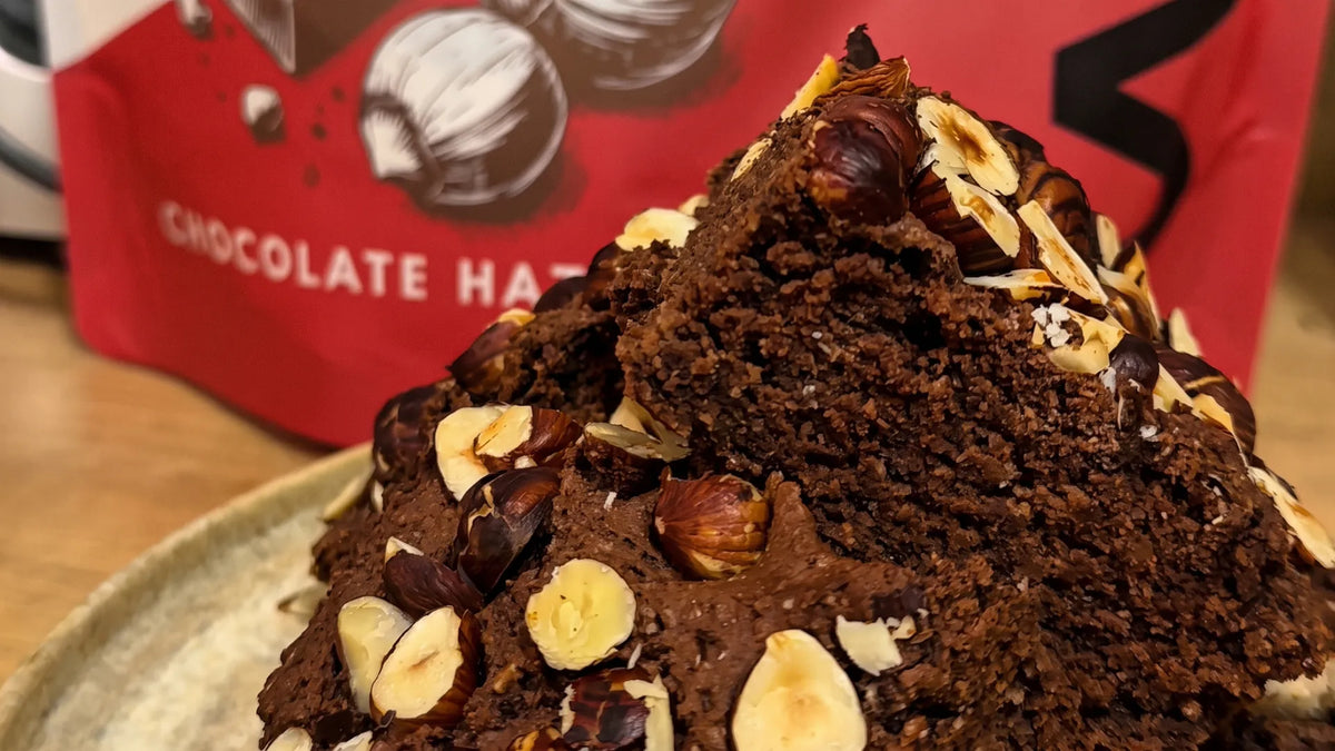 Fudgy vegan protein brownies topped with roasted hazelnuts, in front of a WAM Protein Chocolate Hazelnut pouch