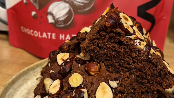 Fudgy vegan protein brownies topped with roasted hazelnuts, in front of a WAM Protein Chocolate Hazelnut pouch