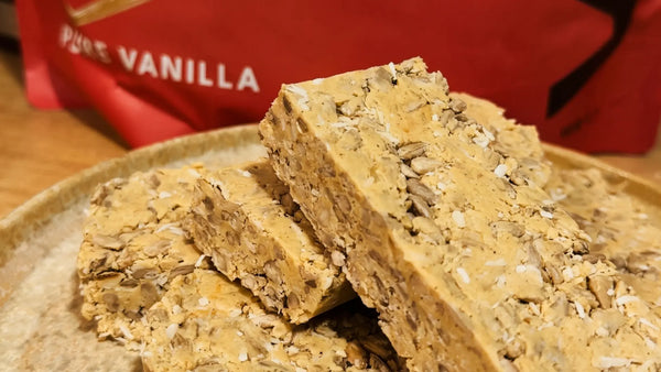 Vanilla coconut protein bars with sunflower seeds, made without nuts, in front of a WAM Protein Pure Vanilla pouch
