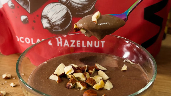 Vegan chocolate protein pudding with chopped hazelnuts in a glass bowl, in front of a WAM Chocolate Hazelnut pouch